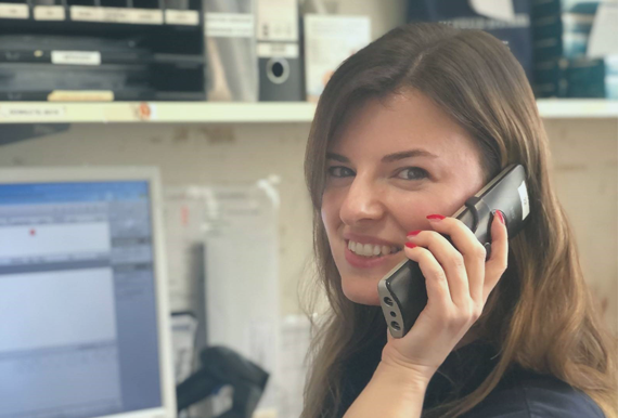 Apothekenmitarbeiterin schaut in die Kamera und lächelt, sie hält ein Telefon am Ohr